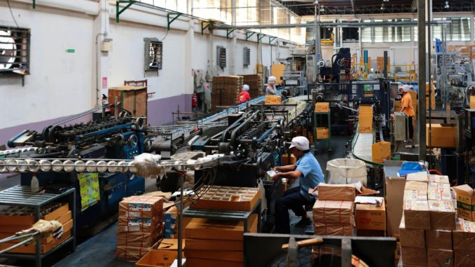 Mitarbeitende in Industriehalle mit Laufbändern und anderen lärmenden Maschinen ohne Lärmschutzmaßnahmen